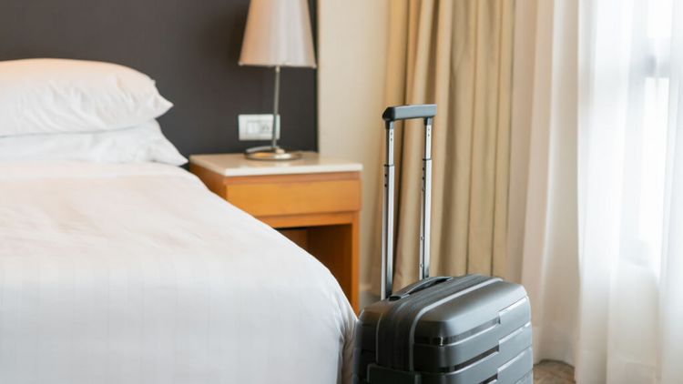 ein schwarzer Rollkoffer wartet vor einem frisch gemachten Bett in einem Hotelzimmer